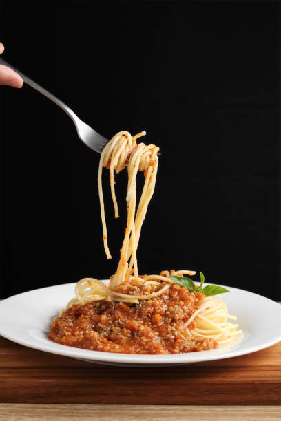 istock Hand holding a fork with spaghetti and red sauce in white dish on wooden cutting board 1324220369