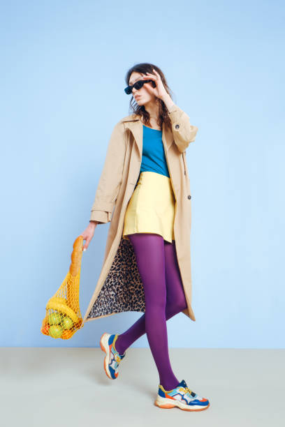 A young fashion girl in a beige trench, yellow short skirt, purple tights and sunglasses holds a mesh reusable eco bag with food. A young fashion girl in a beige trench, yellow short skirt, purple tights and sunglasses holds a mesh reusable eco bag with food. Blue background. pantyhose stock pictures, royalty-free photos & images