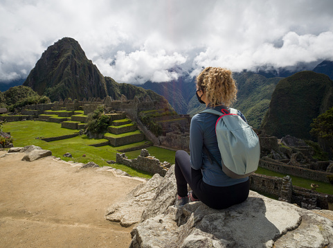 Female tourist sitting on a rock at Machu Picchu and admiring the beautiful view and wearing a facemask - travel destinations concepts