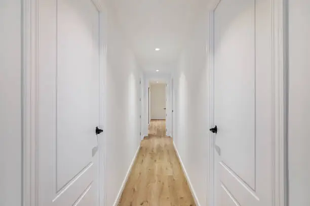 Photo of Interior of long narrow hallway with closed doors, wooden floor and white walls in apartment designed in minimal style