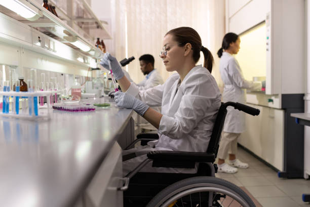 Side view of female lab worker in wheelchair experimenting with fluids Side view of female lab worker in wheelchair experimenting with fluids in flasks laboratory chemist scientist medical research stock pictures, royalty-free photos & images