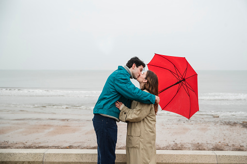 Side view of a young couple under an umbrella outdoors at the beach in a rainy Whitley Bay, they are kissing each other.