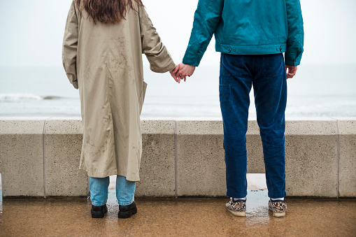Rear view of an unrecognisable young couple holding hands while standing on a walkway near a the beach in rainy Whitley Bay.