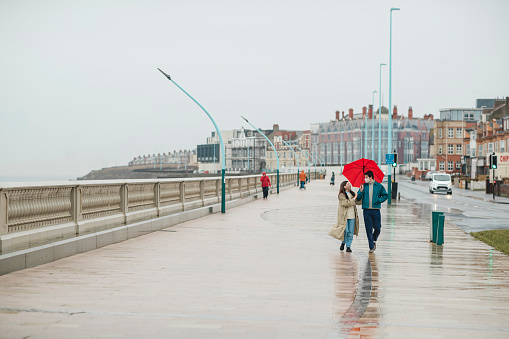 A young couple walking down a promenade at the beach in rainy Whitley Bay, they are wearing protective face masks to reduce contact during the COVID-19 pandemic. The man is holding an umbrella over them while they walk and talk.