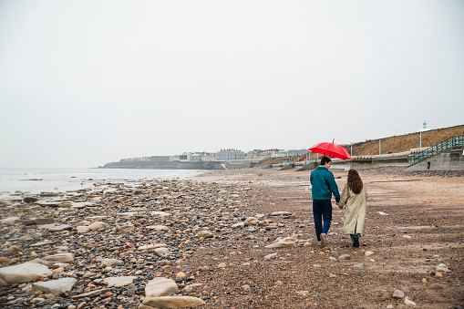 Rear view of a young couple walking along the beach in rainy Whitley Bay, they are holding hands while they walk. The man is holding an umbrella over them.