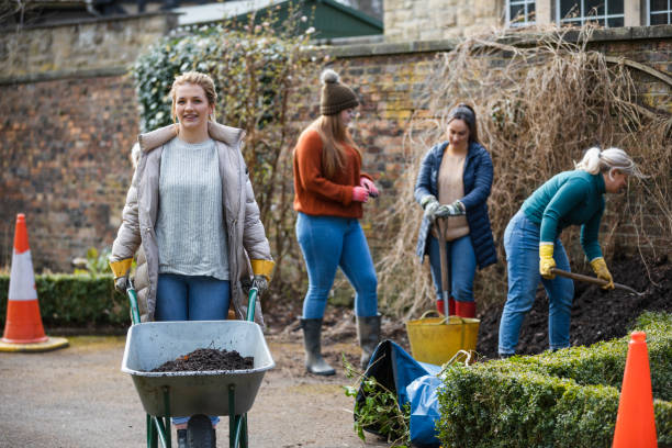 Helping In The Community A front view of a young woman pushing a wheelbarrow with a content look on her face as she helps with a group of women as they work on a community garden outreach project in Hexham in the North East of England. community garden sign stock pictures, royalty-free photos & images