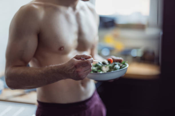 entrenamiento en casa: fit young man comiendo ensalada después del entrenamiento - muscular build fotografías e imágenes de stock