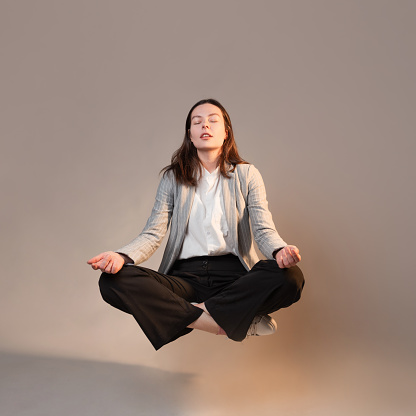 Meditation and stress control at work, a young woman in a business suit, relaxed in the lotus position, levitating in the air, photo connage on a gray background