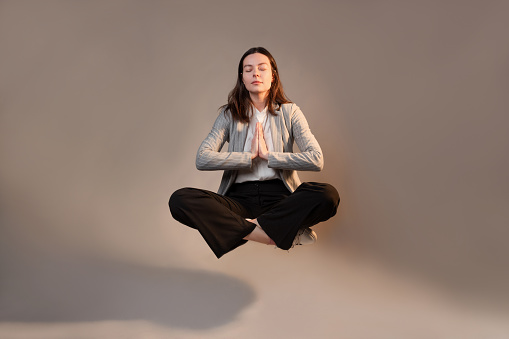 Meditation and stress control at work, a young woman in a business suit, relaxed in the lotus position, levitating in the air, photo connage on a gray background