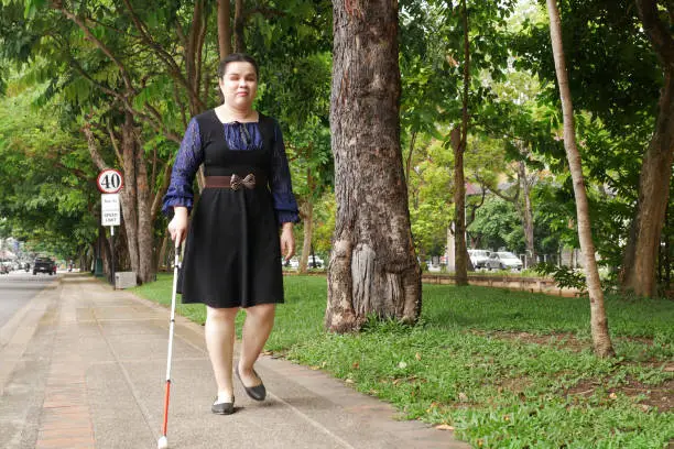 Asian blind person woman walking on sidewalk with a long white cane a mobility tool used to detect objects in the path, also helpful for onlookers in identifying the user as blind or vision disability