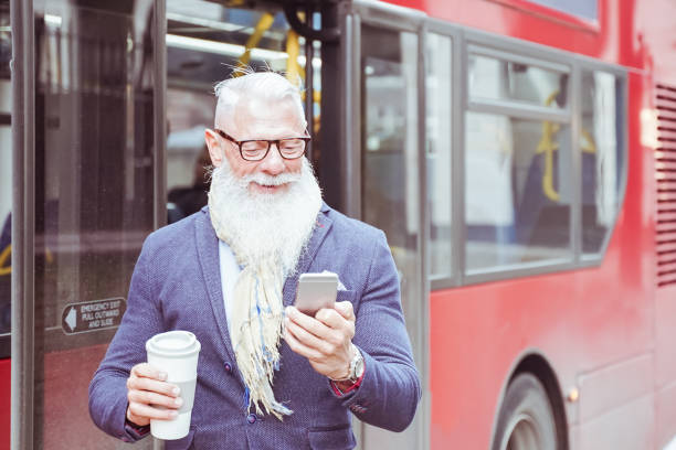 szczęśliwy starszy biznesmen pije kawę i odchodzi z pracy. człowiek chodzący w deszczowy dzień w środku jesieni. człowiek czeka z autobusu i szuka smartfona. koncepcja pracy i stylu życia mody. - senior adult outdoors physical activity mode of transport zdjęcia i obrazy z banku zdjęć