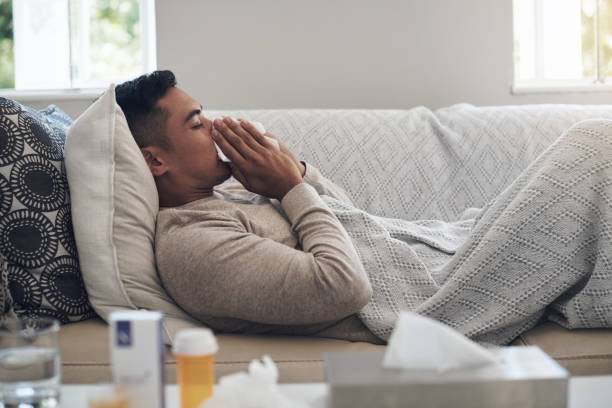 shot of a young man blowing his nose while feeling sick at home - resfriado imagens e fotografias de stock