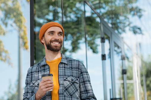 Handsome smiling man holding take away coffee cup looking away standing outdoors. Coffee break concept