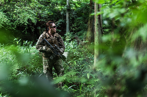 An Asian ethnicity Military soldier in Multicam in a woodland area with a M4 carbine rifle in custom camouflage, ACOG sight, lasers and tactical light.