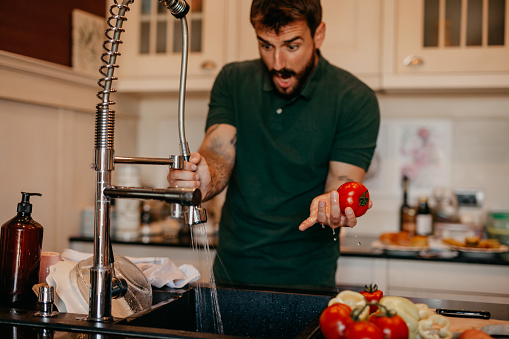 Handsome shocked man is about to wash a tomato and other vegetables on the sink, water is blowing all around the kitchen.