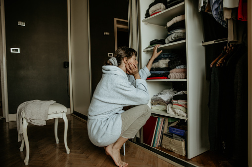 A spontaneous photo of a cute young adult mom still waking up while being in her bathrobe in a squatting position is front of her wardrobe thinking what she has to work on and wear today