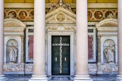 Rome, Italy, June 15 -- The main door under the colonnade of the Basilica of Saint Paul Outside the Walls (San Paolo fuori le Mura) in the Ostiense district of Rome. Built in 324 A.D. at the behest of the Roman emperor Constantine on the tomb of the Apostle Paul, the Basilica was enlarged to its present form starting from 386 AD. by Emperor Theodosius I and was consecrated in 402 AD. by Pope Innocent I. This sacred place for Christianity was declared a World Heritage Site by Unesco in 1980. The Basilica of Saint Paul is one of the four ancient major basilicas of Rome, along with the basilicas of Saint John in the Lateran, Saint Peter's, and Saint Mary Major. Image in high definition format.