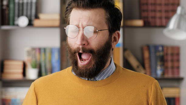 Man yawns. Tired sad bearded man in glasses in office or apartment room yawns, he is bored or wants to sleep. Close up Man yawns. Tired sad bearded man in glasses in office or apartment room yawns, he is bored or wants to sleep. Close up view irony stock pictures, royalty-free photos & images