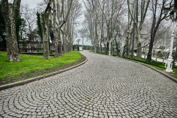 Cobblestone way and path in Gulhane public park with white street lamps and withered and dried trees and green grass during overcast sky and rain and bosporus background.