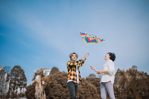 asian chinese teenage boy and his grandmother playing kite in public park during sunny day morning