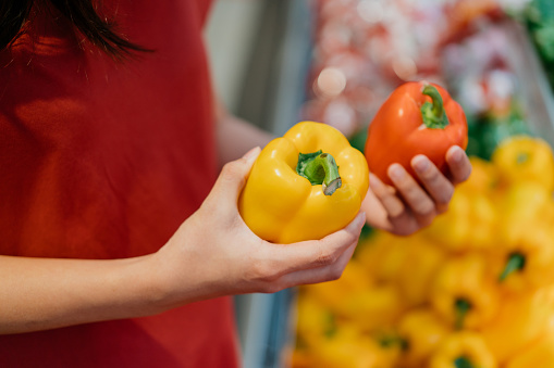 Image of an Asian Chinese woman shopping for fresh bell peppers in supermarket. Woman holding red and yellow bell pepper, deciding which one to buy.