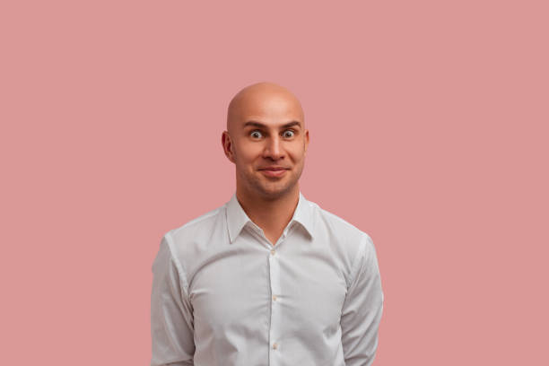 And what next. Pleasantly shocked bald guy in white shirt holds hands behind back. Keeps eyes widely opened, looks what happens next. Isolated over pink background