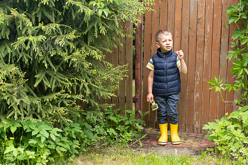 Little boy in rubber boots stands against the background of a wooden rustic fence.