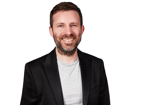 Portrait of a man with dark hair and a beard in his 40s wearing a black jacket and gray t-shirt. High resolution photographed against a white background as a cutout