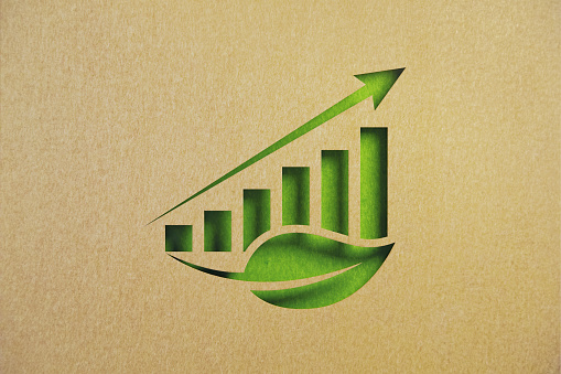 Cut out leaf shapes made of recycled paper intersect with a bar graph on green background. Horizontal composition with copy space. Sustainability concept.