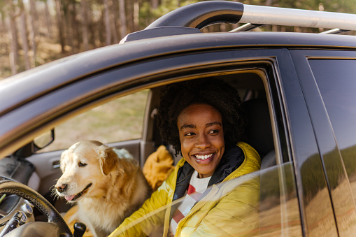 Photo of a young smiling woman driving a car. Her dog is sitting on a passenger seat