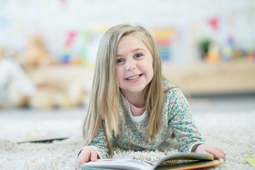 A girl smiles at the camera while reading a book on the carpet.