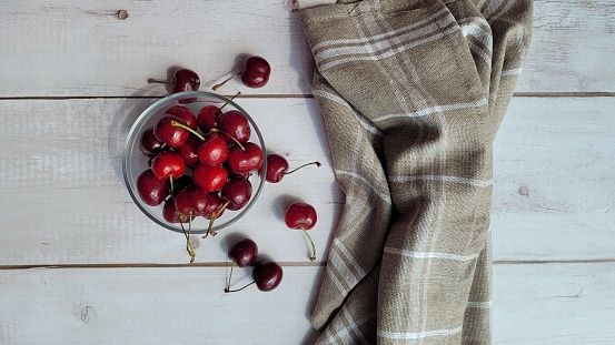 Sweet red cherries in glass bowl, on white wooden old table outdoors and brown napkin next to the bowl. Copy space available. Food on table in sunny day. Unedited image. Eating fruits outdoors on rustic wooden old table.