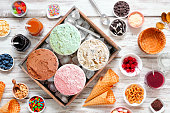 Summer ice cream buffet table with a variety of flavors and sweet toppings. Overhead view on rustic white wood.