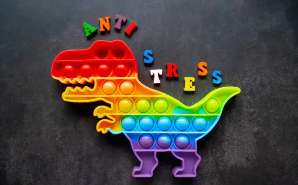 Photo of Toy pop it dinosaur rainbow colors on a black background with multicolored letters and the inscription-Anti-stress on the back