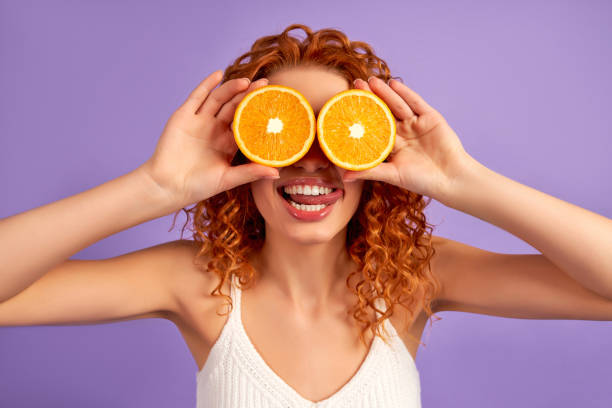 Redhead girl on background Cute redhead girl with curls holds halves of oranges instead of eyes isolated on purple background. Proper nutrition. Healthy lifestyle. oranges with faces stock pictures, royalty-free photos & images