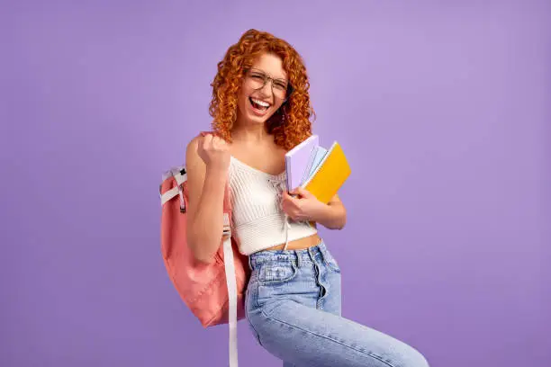 Cute redhead curly girl student teenager with a backpack holds notepads and notebooks and shows the gesture yes as a winner isolated on a purple background.