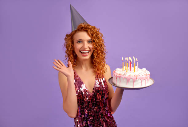 Redhead girl on background Cute red-haired woman with curls in a shiny dress and a party cap is holding a cake isolated on a purple background. The concept of a party, celebration, birthday. woman birthday cake stock pictures, royalty-free photos & images