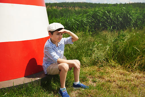 Adorable child boy sitting on the background of a bright lighthouse and looking into the distance. Concept of spending time outdoor.