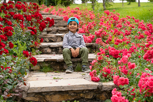 Botanik bahçesinde, güllerin arasından uzanan merdivende oturan çocuk portresi. kameraya bakıp gülümsüyor. kırmızı ve pempe renkli güller full frame makine ile çekilmiştir.