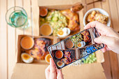 Personal perspective human hand taking photo using smart phone on table top view malaysian food nasi kerabu, nasi ulam and ayam percik in recycled paper container with sauce