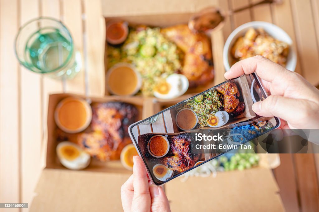 Personal perspective human hand taking photo using smart phone on table top view malaysian food nasi kerabu, nasi ulam and ayam percik in recycled paper container with sauce Take Out Food Stock Photo