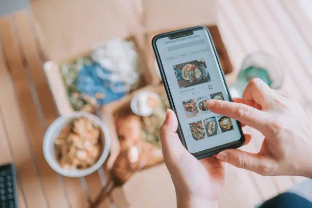 Photo of high angle view Close up Asian woman using meal delivery service ordering food online with mobile app on smartphone in the living room at a cozy home
