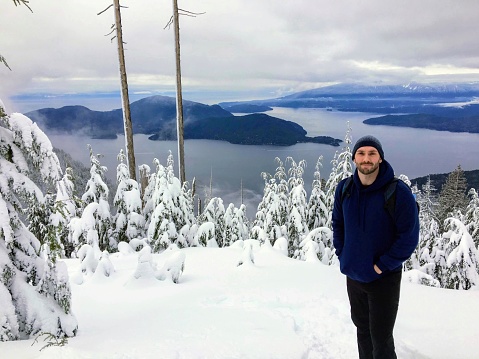 A handsome man posing for a photo dressed in a winter coat and toque, with the forest and ocean in the background, along the bowen island lookout trail, on cypress mountain, british columbia, canada.