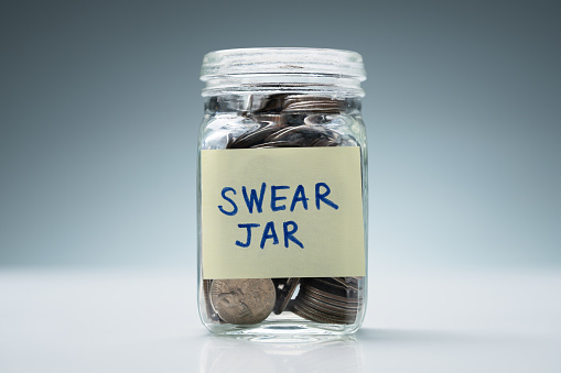 Close-up Of A Glass Jar With Swear Jar Text Filled With Coins Against Grey Background