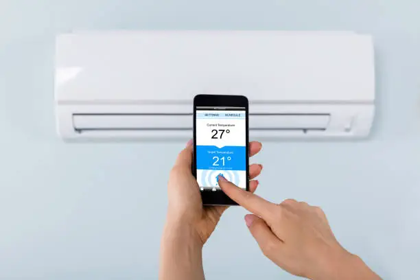 Close-up Of Person Hand Setting Temperature Of Air Conditioner Using Mobilephone At Home