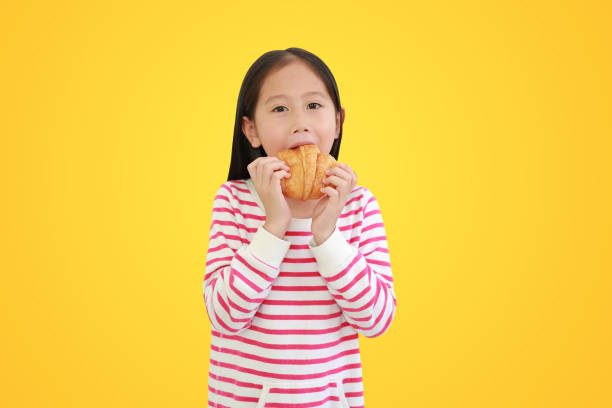 Asian little child girl eating croissant isolated on yellow background with clipping path
