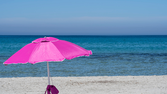 Isolated fuchsia or pink beach umbrella. Blue sky. Relaxing context. Summer holidays at the sea. General contest and location. Turquoise sea and white beach in the background