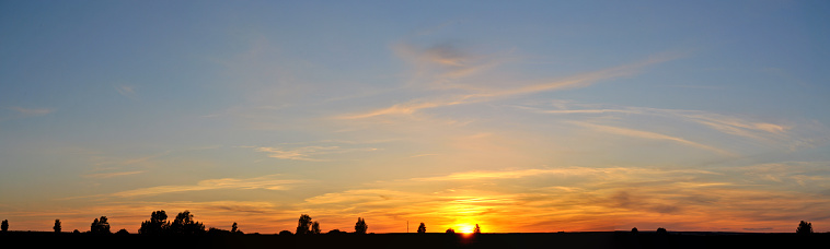 A panoramic view of a bright summer sunset in the field and clouds illuminated by the setting sun.The silhouettes of several trees are visible on the horizon.