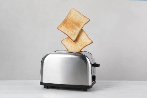 Photo of Bread slices popping up from modern toaster on white wooden table