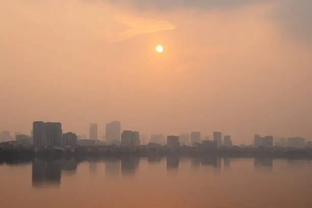 sunset over westlake, hanoi with buildings reflected in the water and air pollution causing an orange colour